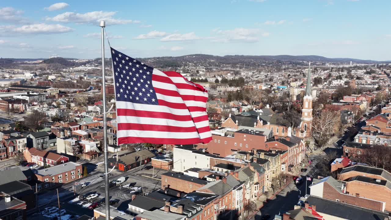 Veduta aerea di York, Pennsylvania, con una grande bandiera americana che sventola sopra edifici storici. Paesaggio urbano tranquillo con chiese, tetti e dolci colline sotto la luce chiara del giorno. Ripresa all'indietro.