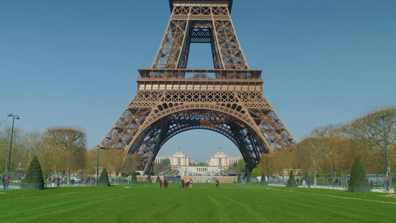 Prato verde alla base della Torre Eiffel con persone che camminano sullo sfondo, ripresa a mano. Vista panoramica della struttura in acciaio simbolo di Parigi con turisti sull'erba alla luce del giorno