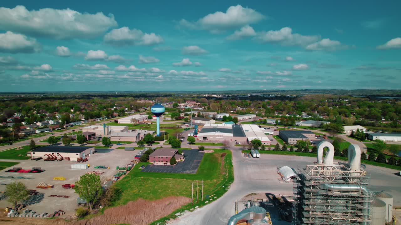 Elevated drone view of De Pere, Wisconsin, showing an industrial facility with silos and a scenic town skyline beneath scattered clouds.
