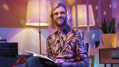 Young-stylish-caucasian-man-with-long-hair-in-glasses-sitting-on-a-chair-and-reading-a-book-in-a-retro-room-at-home