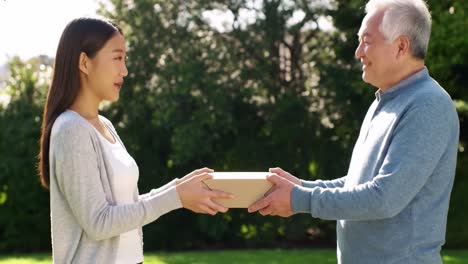 a young woman hands a small package to an elderly man in a sunny outdoor setting. they smile at each other, enjoying the pleasant atmosphere and the act of kindness.