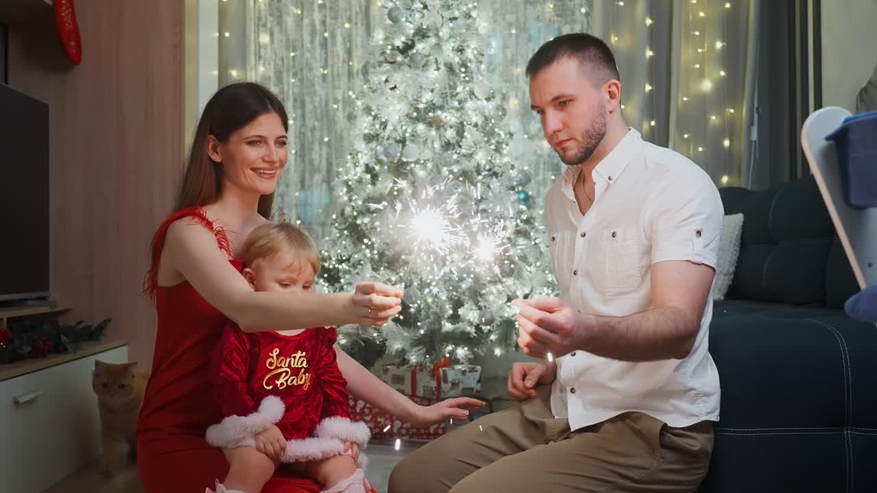 Happy family, mother, father and baby daughter, wearing red, smiling and playing with sparklers, celebrating Christmas eve in the living room