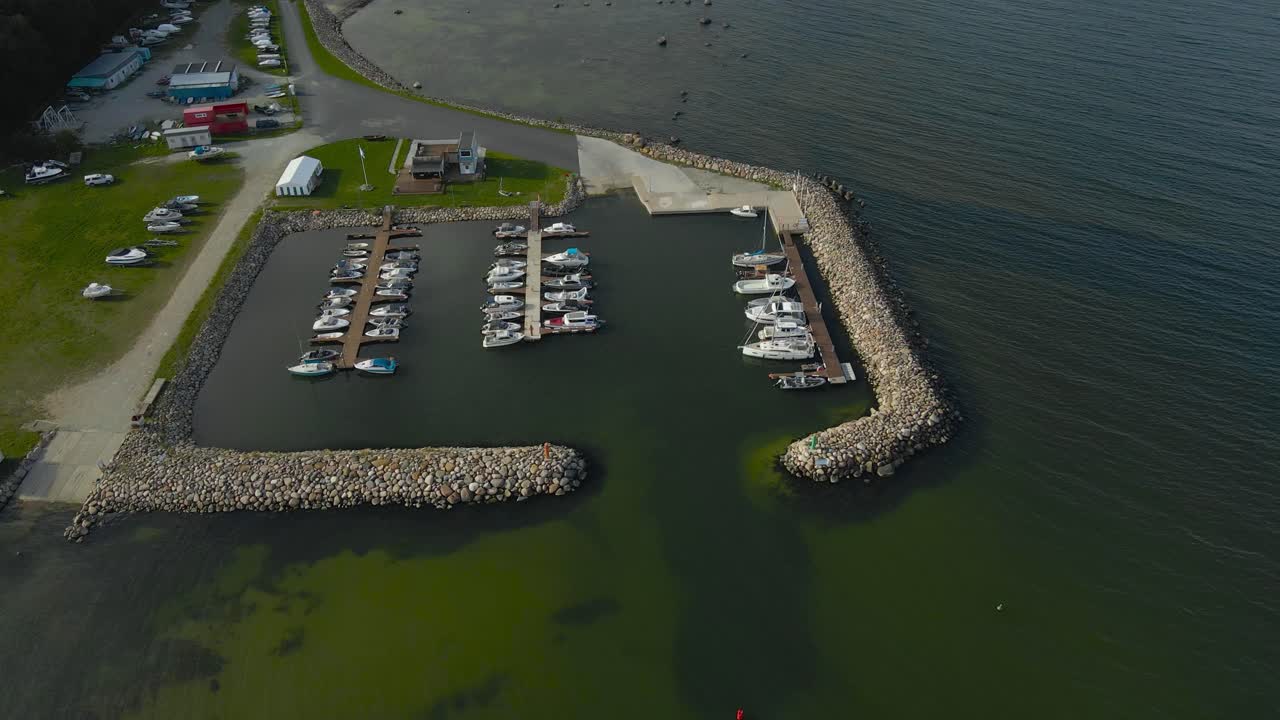 Aerial drone footage orbiting and flying around a small luxury sailboat and yacht harbor where the boats are docked on the rocky ocea sea pier. Shallow sea has sand visible on the bottom, green water