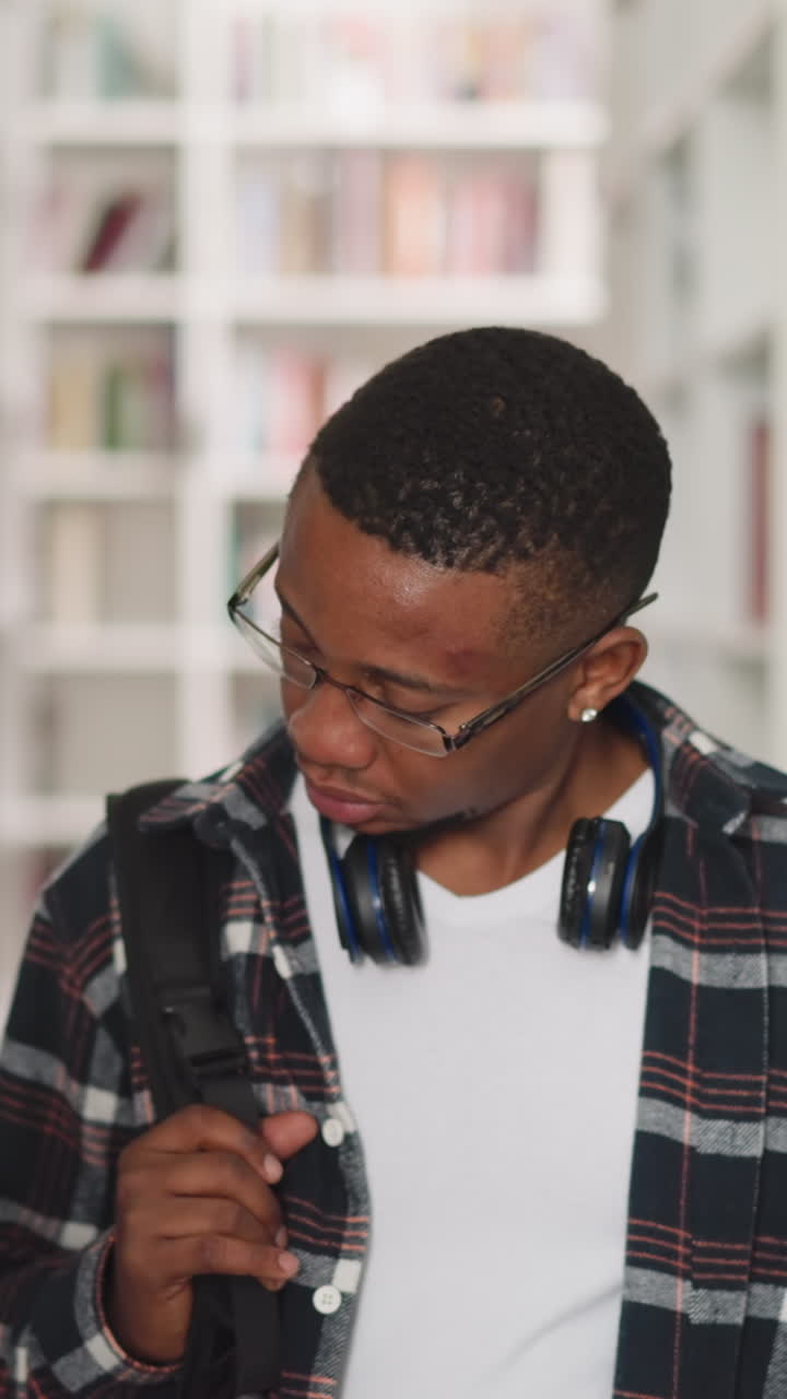 Black man looks around in public library. African American visitor with glasses and headphones chooses books in store. Education materials storage
