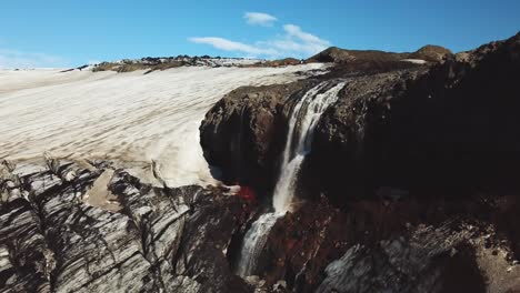 Aerial-landscape-view-of-a-glacial-river-flowing-down-a-waterfall-next-to-an-icelandic-glacier,-on-a-sunny-day