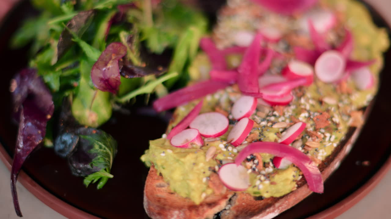 Close up of an avocado toast with seeds, radish, red onion and a side salad on a black plate