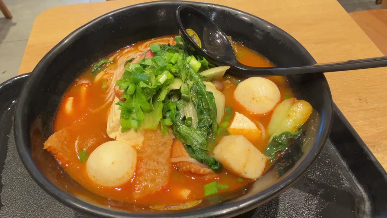 A steaming bowl of spicy hotpot with fish balls, tofu, leafy greens, and noodles in red broth, garnished with chopped scallions, served with a black spoon.