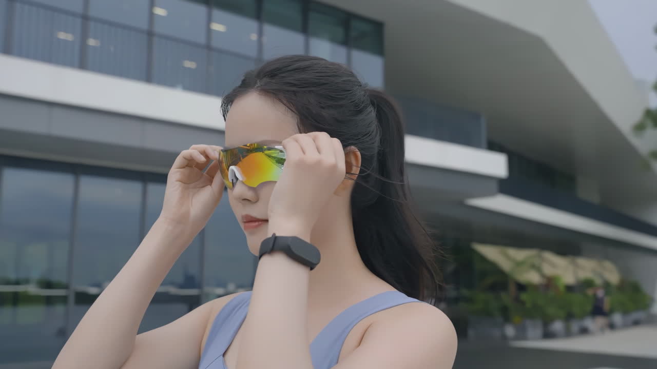 Athletic runner woman in sports clothes puts on glasses prepares for running exercise. Athlete female in fitness outfit and protective eyeglasses standing on park preparing jogging outdoors