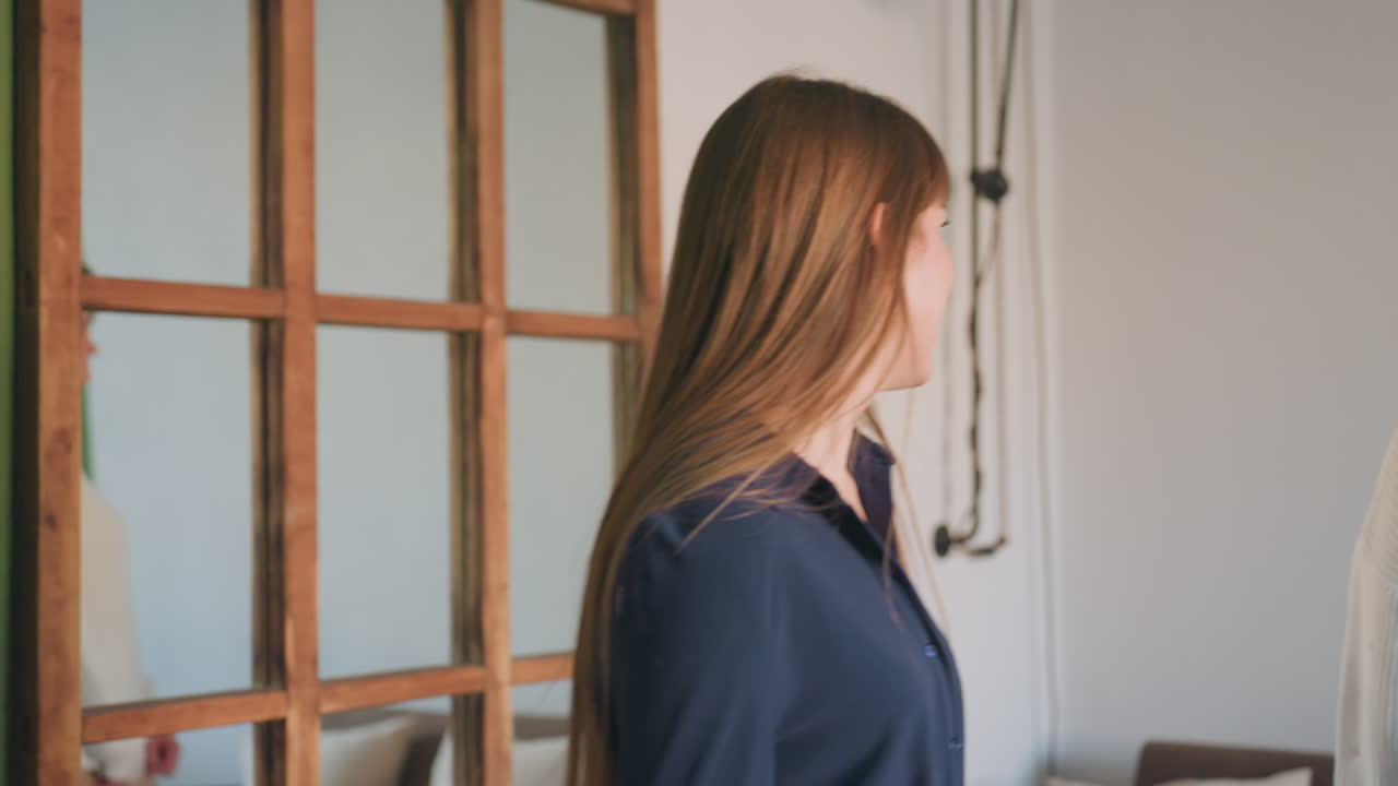 Practitioner with bold smile welcomes participant to session with eye contact and warm energy, both preparing to sit and engage in supportive conversation in bright, calming therapy environment