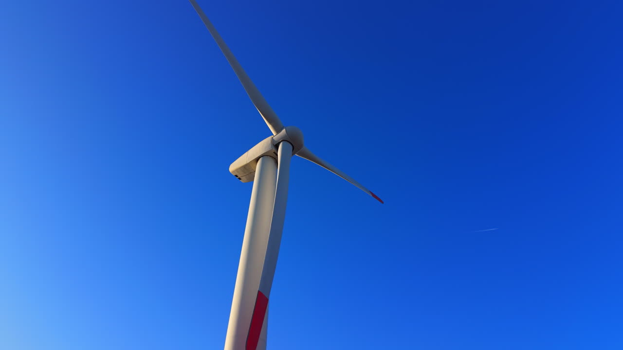 Turbina eolica bianca con tre pale su uno sfondo di cielo azzurro intenso e uniforme durante il giorno. Vista ravvicinata di una singola turbina eolica contro un cielo sereno.