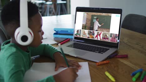 Schoolboy-using-laptop-for-online-lesson-at-home,-with-diverse-teacher-and-class-on-screen