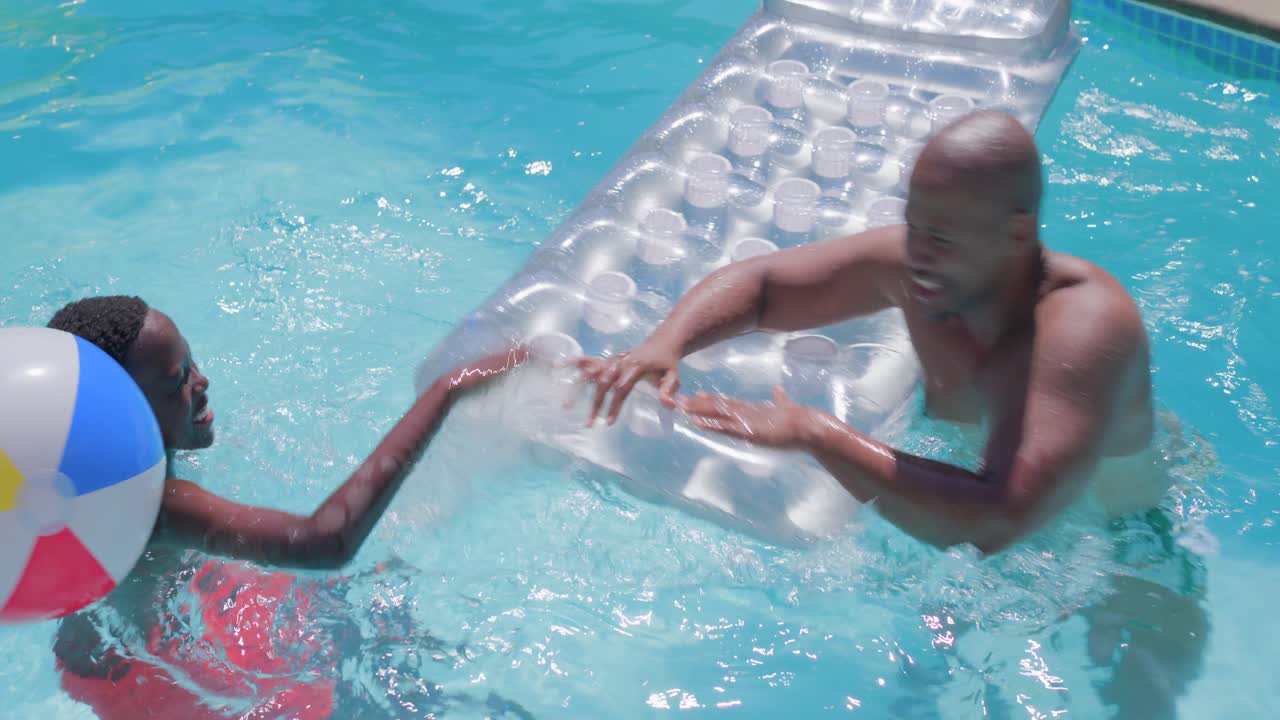 Splashing dad in green trunks and red-trunked son pushing silver float and ball yard pool playfully