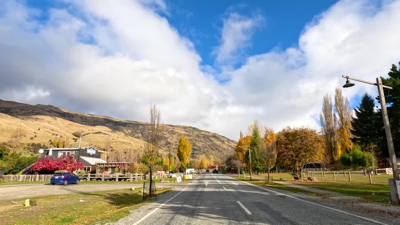 A picturesque drive through Wanaka, New Zealand, showcasing vibrant autumn foliage, clear skies, and mountainous terrain under bright daylight