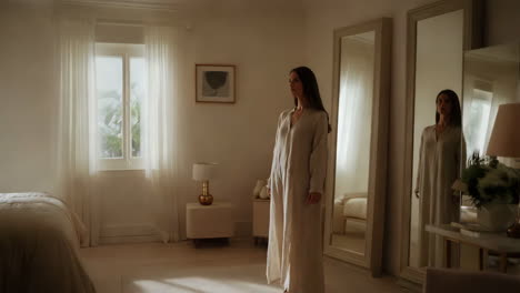 woman in a long robe standing in a well-lit bedroom with a mirror