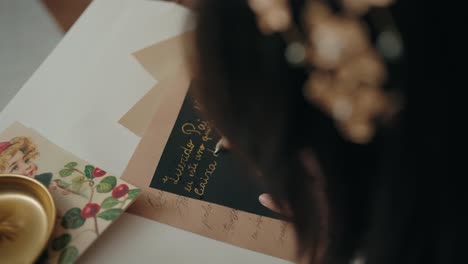 close up of a hand writing a holiday card with a feather quill in gold ink on black paper, with vintage details around