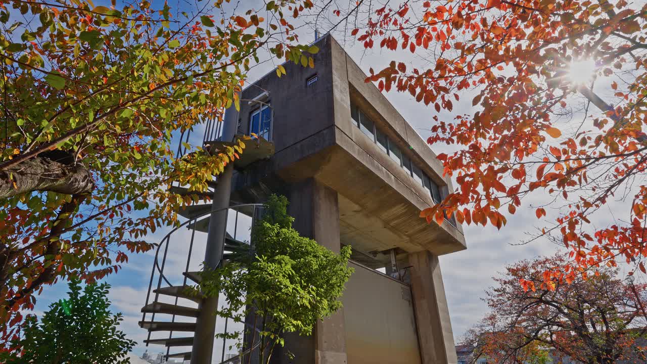 A low-angle view of the concrete pumping station building with an external spiral staircase, framed by bright autumn leaves