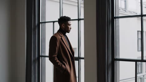 a man in a brown coat stands thoughtfully by a large window