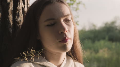 woman with blade of grass in mouth leans on tree trunk. sunlight on face side. young lady has psychological problems feeling anxiety. self-isolation in nature at sunset