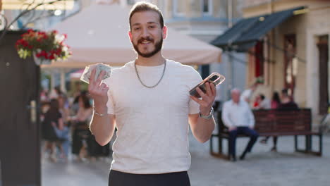 happy young man counting money dollar cash use smartphone calculator app in urban city street