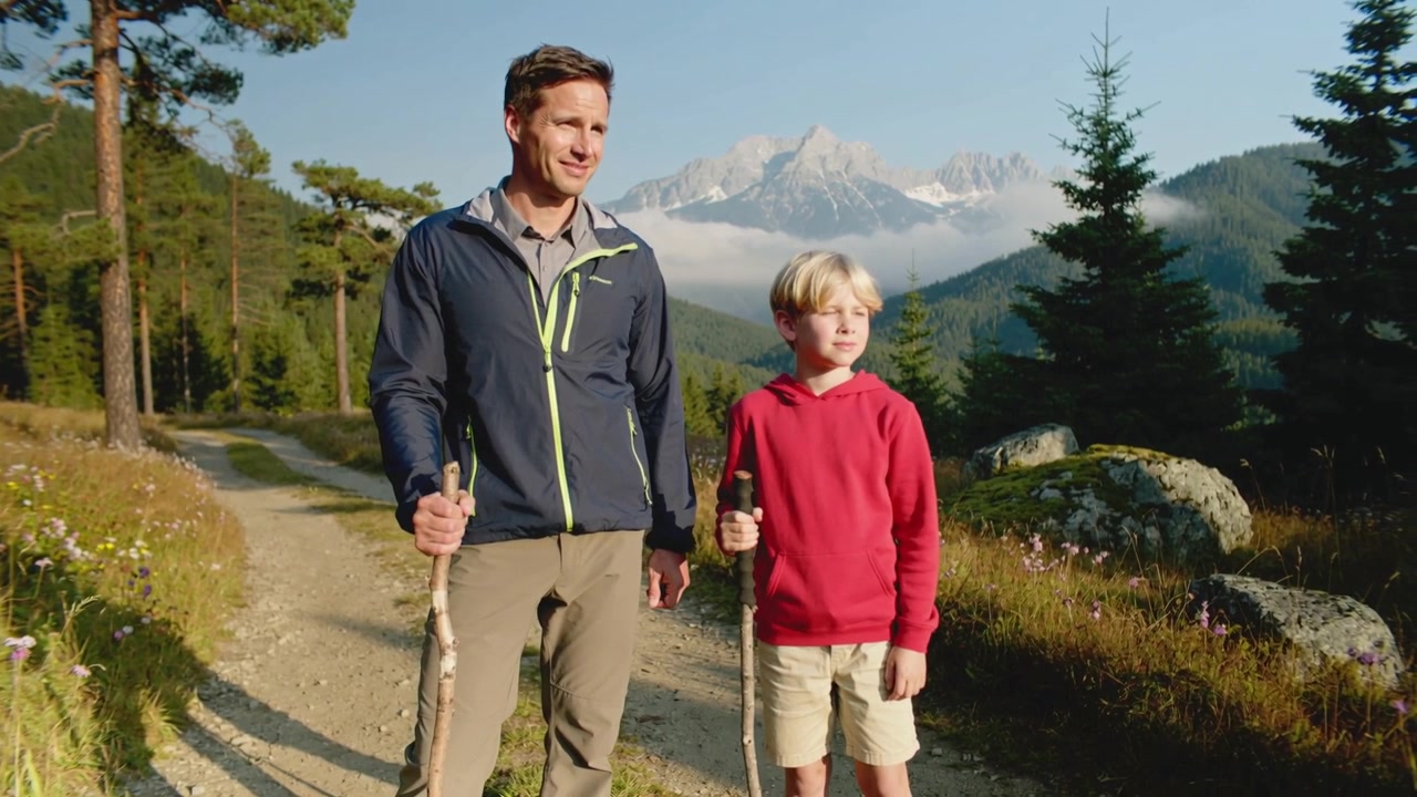 Walking dad dark jacket and son red hoodie pausing, watching mountain view, holding wooden sticks