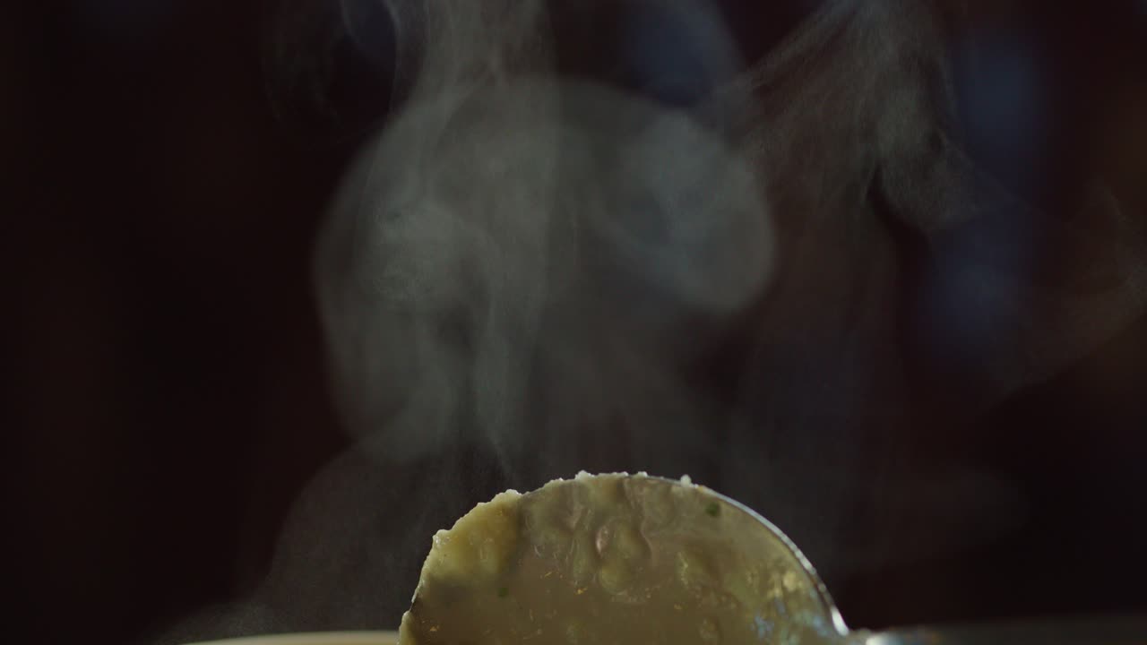 A spoon lifts hot soup, releasing swirling steam in a dark, softly lit environment. Shallow focus and slow camera movement emphasize texture and warmth