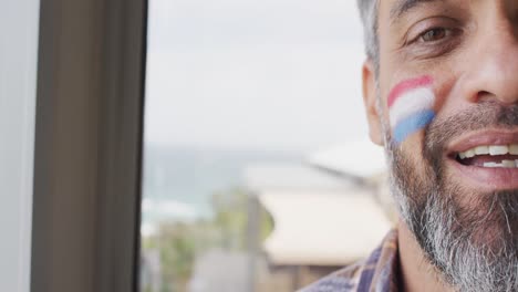 Happy-biracial-man-with-flag-of-netherlands-on-cheek-at-home