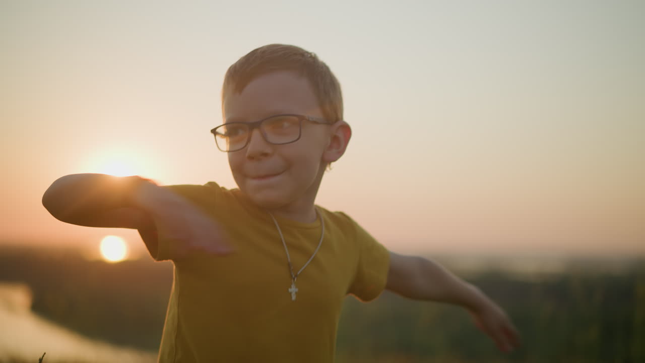 A young boy wearing a yellow shirt smiles shyly while moving his hands in a playful manner, standing in a sunlit grassy field during a serene sunset, capturing the essence of a joyful, carefree moment