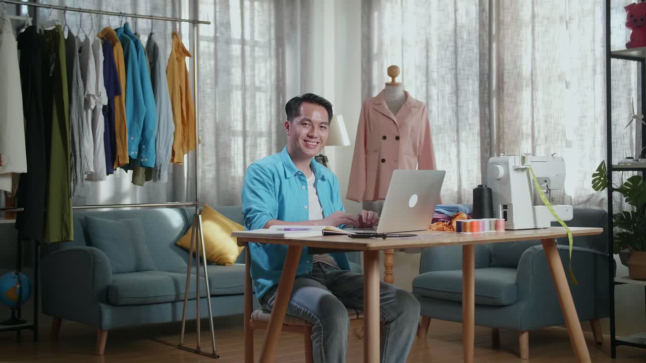 Asian Male Designer Working On A Laptop And Smiling To The Camera While Designing Clothes In The Studio