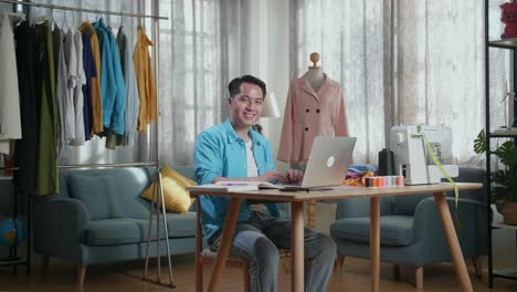 アジアの男性デザイナーがラップトップで作業しスタジオで服をデザインしている間カメラに笑顔を浮かべています