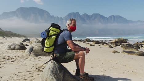 hombre excursionista mayor con máscara facial con mochilas sentado en una roca mientras caminaba por la playa.