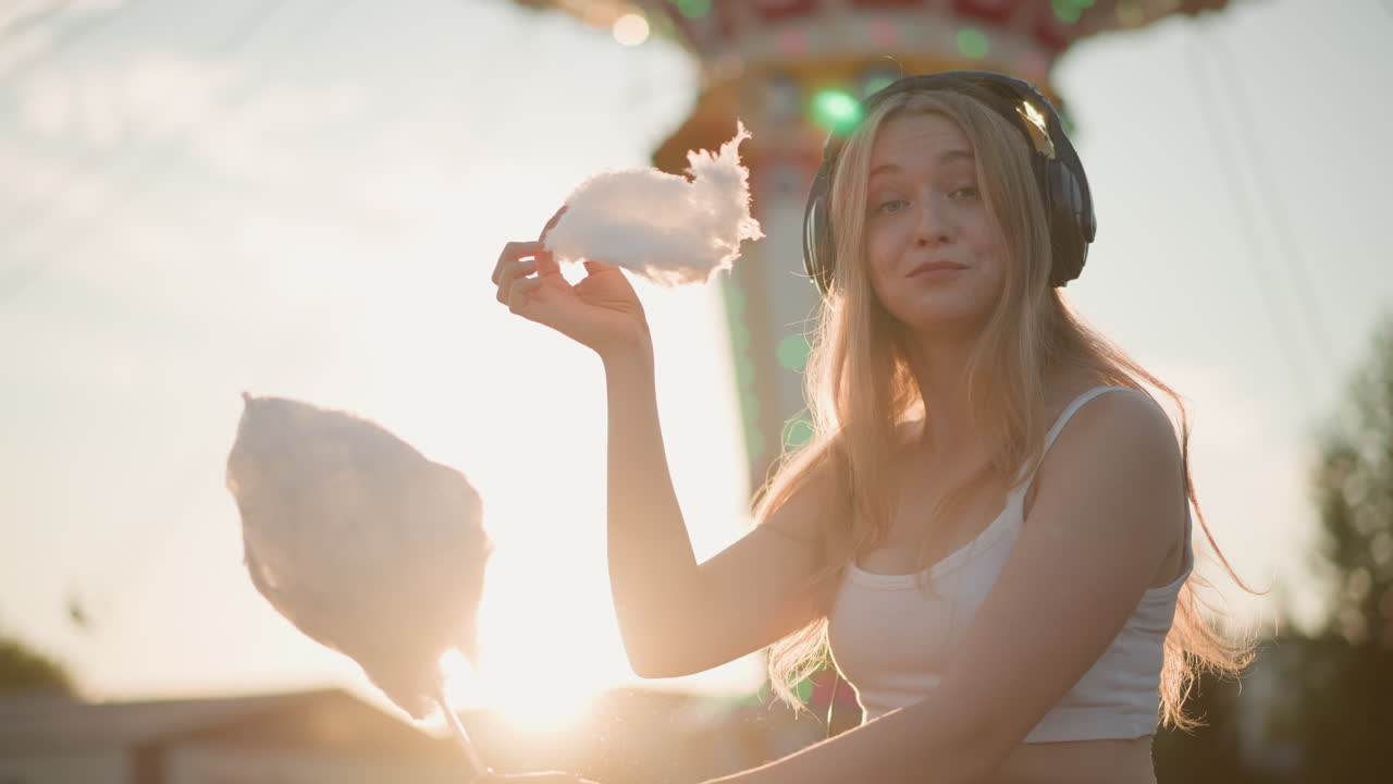 caucasian woman with cotton candy carnival, sunset backlight, fairground tower lights, headphones on, playful gestures, sugar cloud in hand, relaxed seat, sunflare, youthful summer vibe, candid