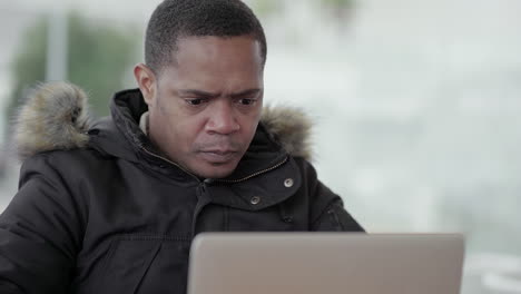 Afro-American-middle-aged-man-in-black-jacket-with-fur-hood-working-on-laptop-outside