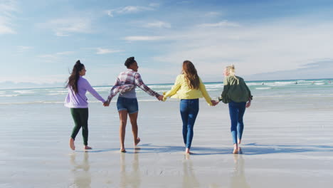 happy group of diverse female friends having fun, walking along beach holding hands