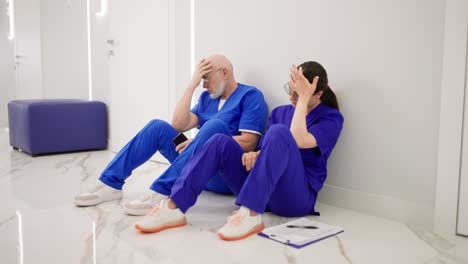 A-confident-and-tired-doctor-an-elderly-man-with-glasses-with-a-white-beard-in-a-blue-uniform-sits-with-his-colleague-a-brunette-girl-on-the-floor-after-a-hard-day-at-work-during-a-break-between-patients-in-a-modern-clinic