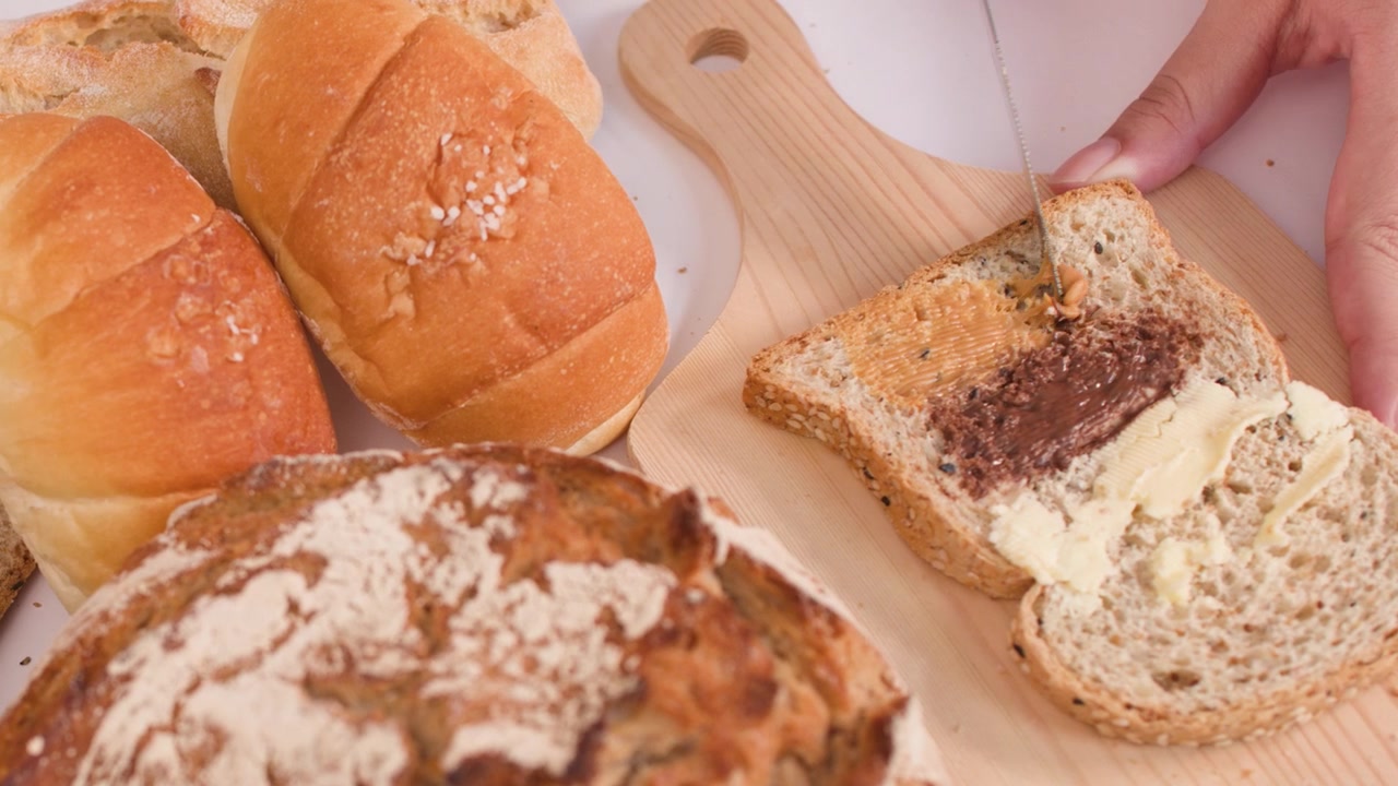 Close-up of hands spreading peanut butter on bread amidst sourdough, baguettes, and croissants.