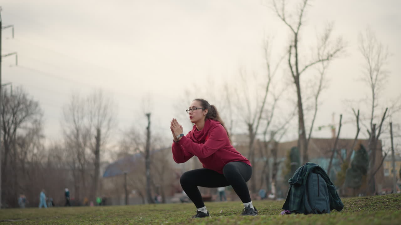 White Woman Warming Up In Park Performing Squats And Side Steps In Red Hoodie By Backpack On Grass Under Overcast Skyline, Focused Expression, Steady Breathing, Dynamic Motion For Cardio And Strength