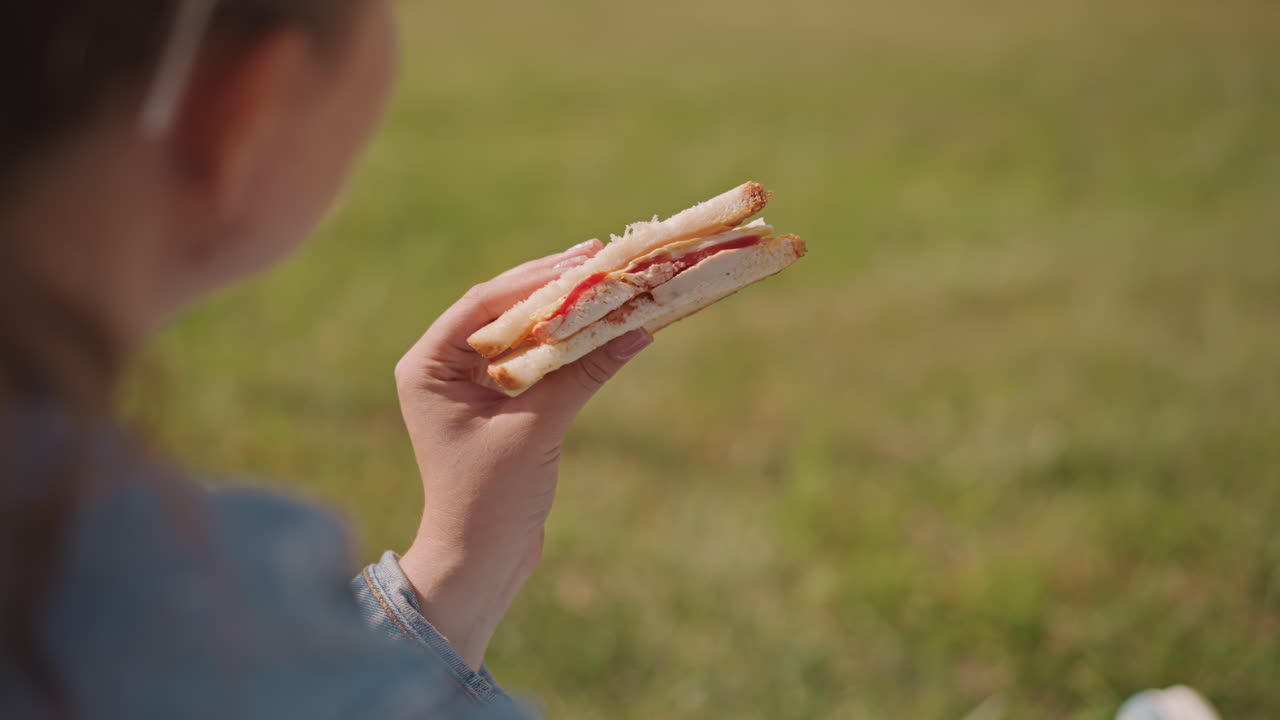 Ragazza caucasica che tiene un panino al parco, luce solare intensa, gesti delle mani animati, primo piano di prosciutto e formaggio a strati, manica della giacca di jeans, routine spontanea del pranzo, energia giovanile, erba verde