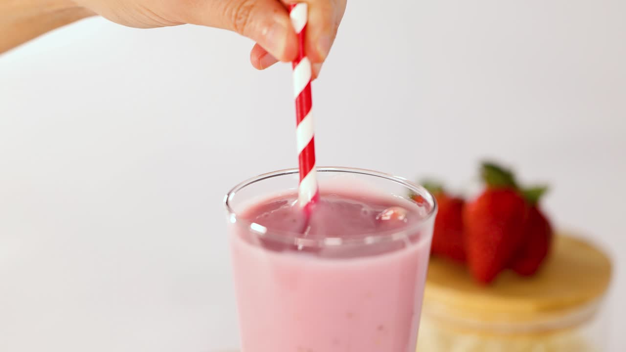 Una mano remueve un cremoso batido de fresa rosa en un vaso, iluminación brillante, poca profundidad de campo, fondo minimalista