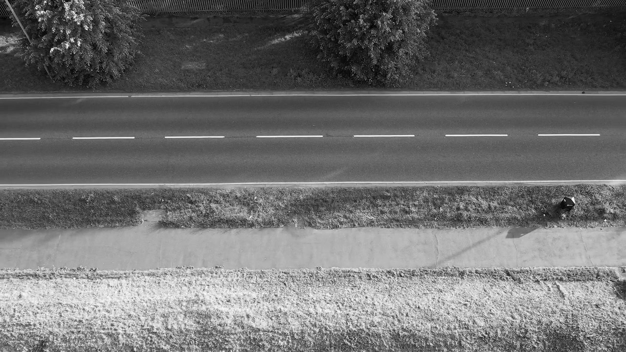 Vertikales Social-Media-Video mit einer Schwarz-Weiß-Drohnenansicht von oben auf eine leere Asphaltstraße, die von Bäumen, Gras und einem Entwässerungsgraben in Marokko begrenzt wird.