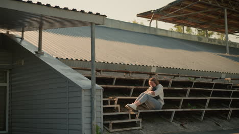 lady seated alone on stadium bleachers in thoughtful pose, hands holding bag, background shows stadium's metal roof, trees, and greenery