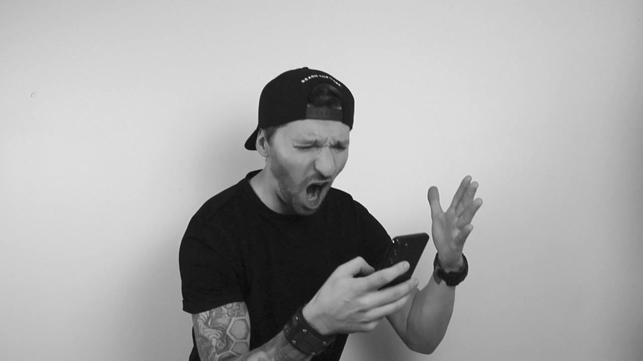 Grayscale footage of a man with black tattoo sleeve watching his phone, getting some bad news, reading something or watching and gets extremely angry. Man scream and yells at the phone, waving hands