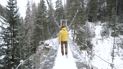 Joven-Caucásico-Con-Chaqueta-Amarilla-De-Invierno-Cruza-Un-Puente-Colgante-En-Un-Bosque-Durante-El-Invierno-Mientras-Nieva