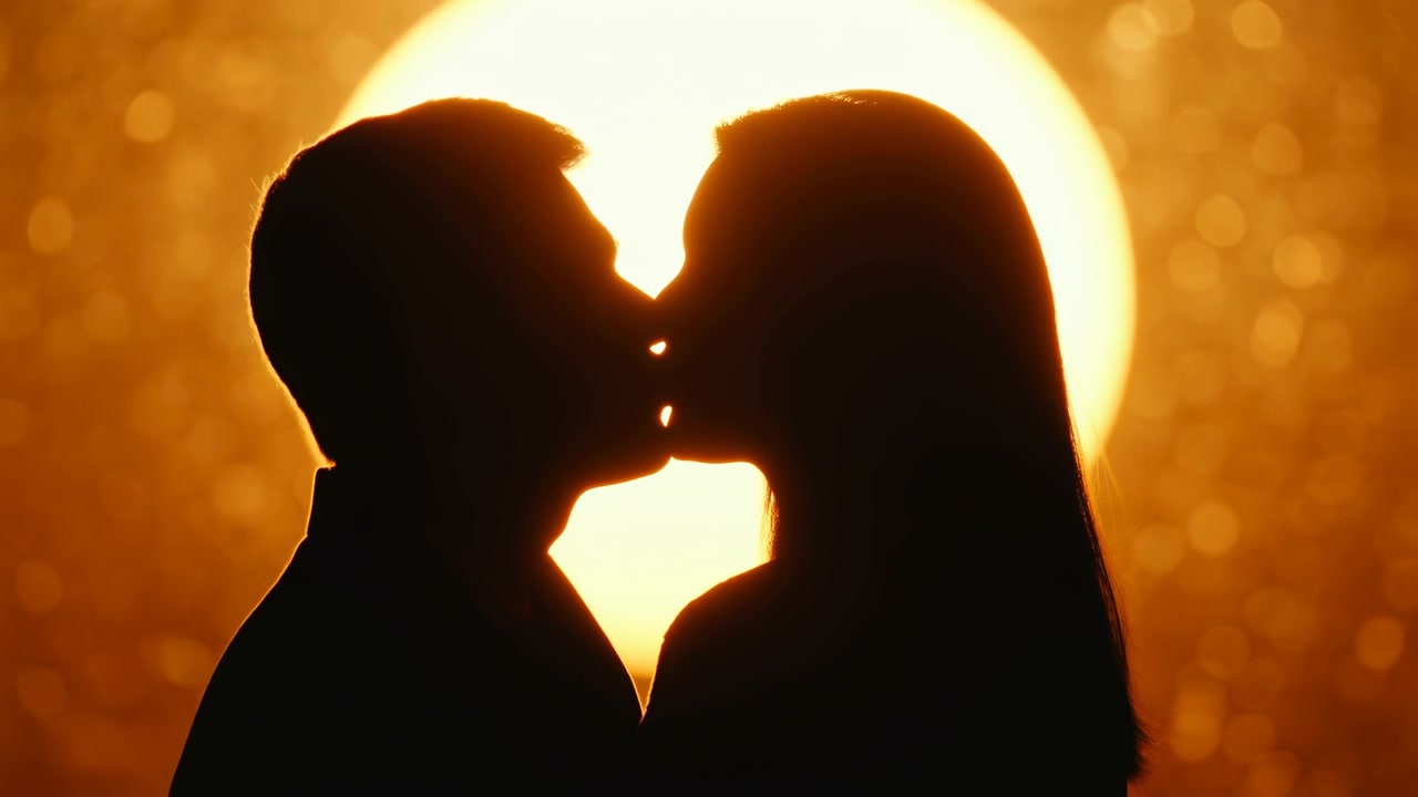 Due figure in silhouette si fronteggiano di profilo ravvicinato mentre si toccano le labbra davanti a un sole basso e luminoso, con un caldo bagliore arancione, sfondo con bokeh morbido, inquadratura stretta e centrata e un'atmosfera calma e intima