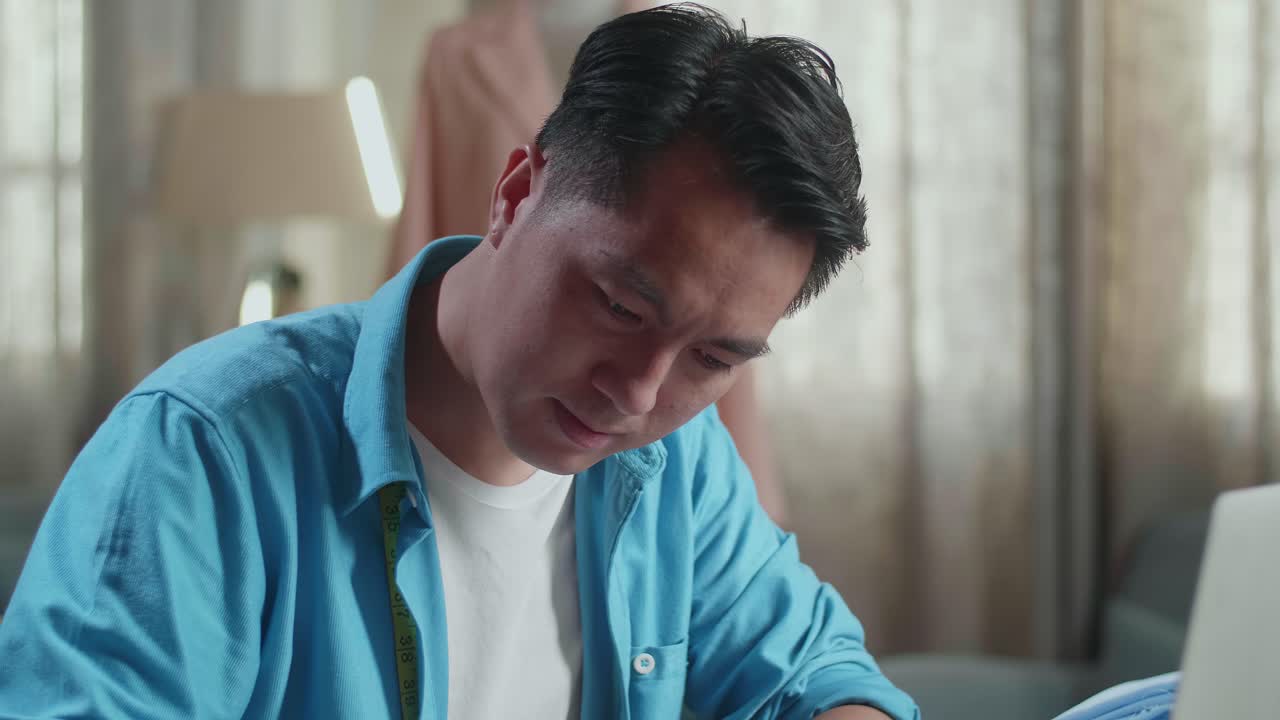 바느질 기계와 함께 아시아 남성 디자이너가 스튜디오에서 노트북 컴퓨터와 함께 작업하는 동안 종이에서 옷을 디자인하는 것을 즐기고 있습니다.