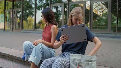 dos mujeres jóvenes sentadas en las escaleras, hablando y usando una computadora portátil