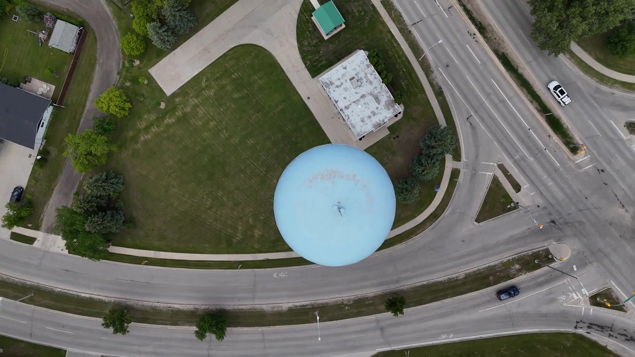 Drone diretamente acima da caixa d'água azul clara em Steinbach, Manitoba, girando para a direita enquanto ascende para revelar as estradas circundantes, carros, casas, árvores, subúrbios e grama verde no verão.