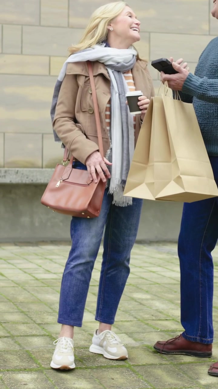 Video verticale: Donna in giacca color cammello che legge il telefono mentre un uomo, con delle borse, condivide il telefono e sorride