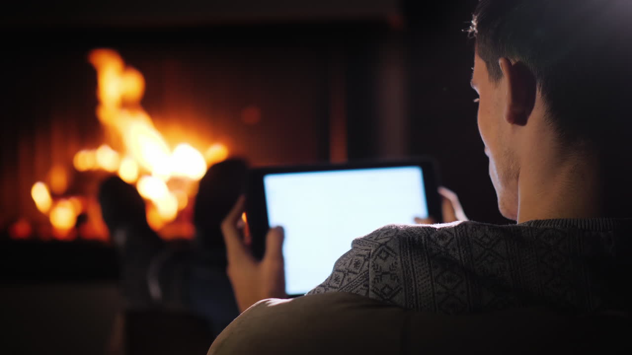 homem confiante com um tablet nas mãos descansando em casa em frente à lareira onde o fogo está