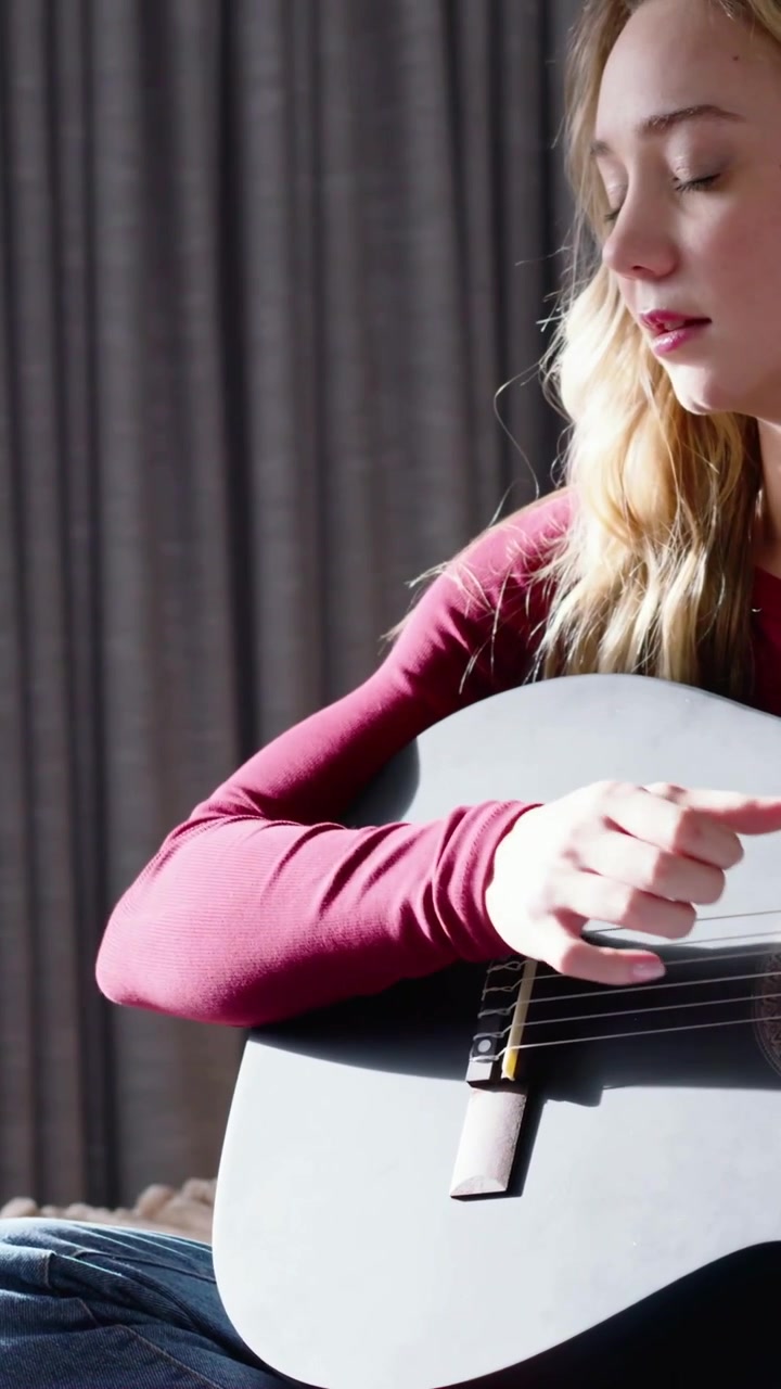 Vertical video: Strumming woman in red top, jeans practicing guitar on sofa, camera panning right