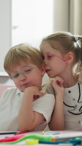 couple of children with painted faces sits at desk with educational equipment touching chins with hands. girl tells boy secret into ear and brother nods in agreement